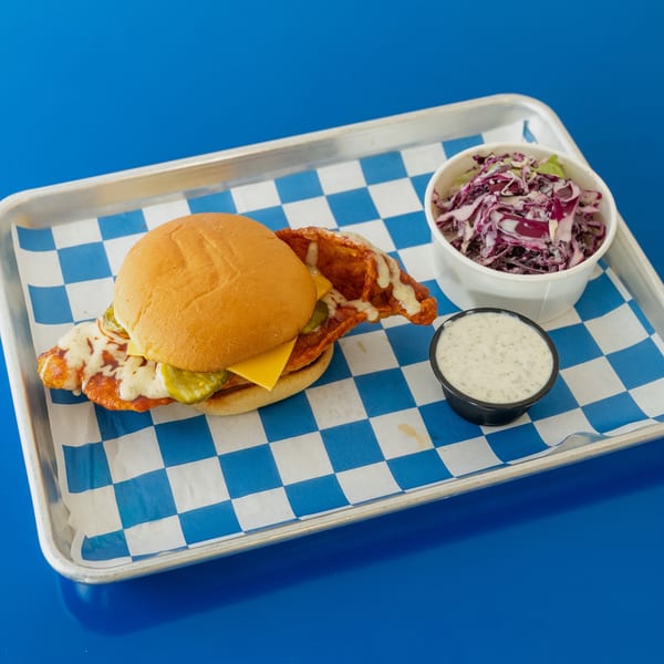 Spicy chicken sandwich with pickles, cheese, and sauce, served with coleslaw and ranch on a tray.