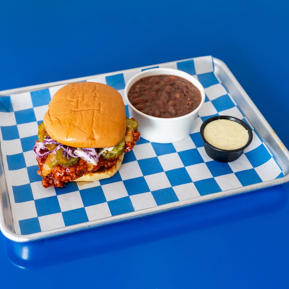 Crispy saucy chicken sandwich with pickles and cabbage slaw, served with beans and creamy dipping sauce on a checkered tray.