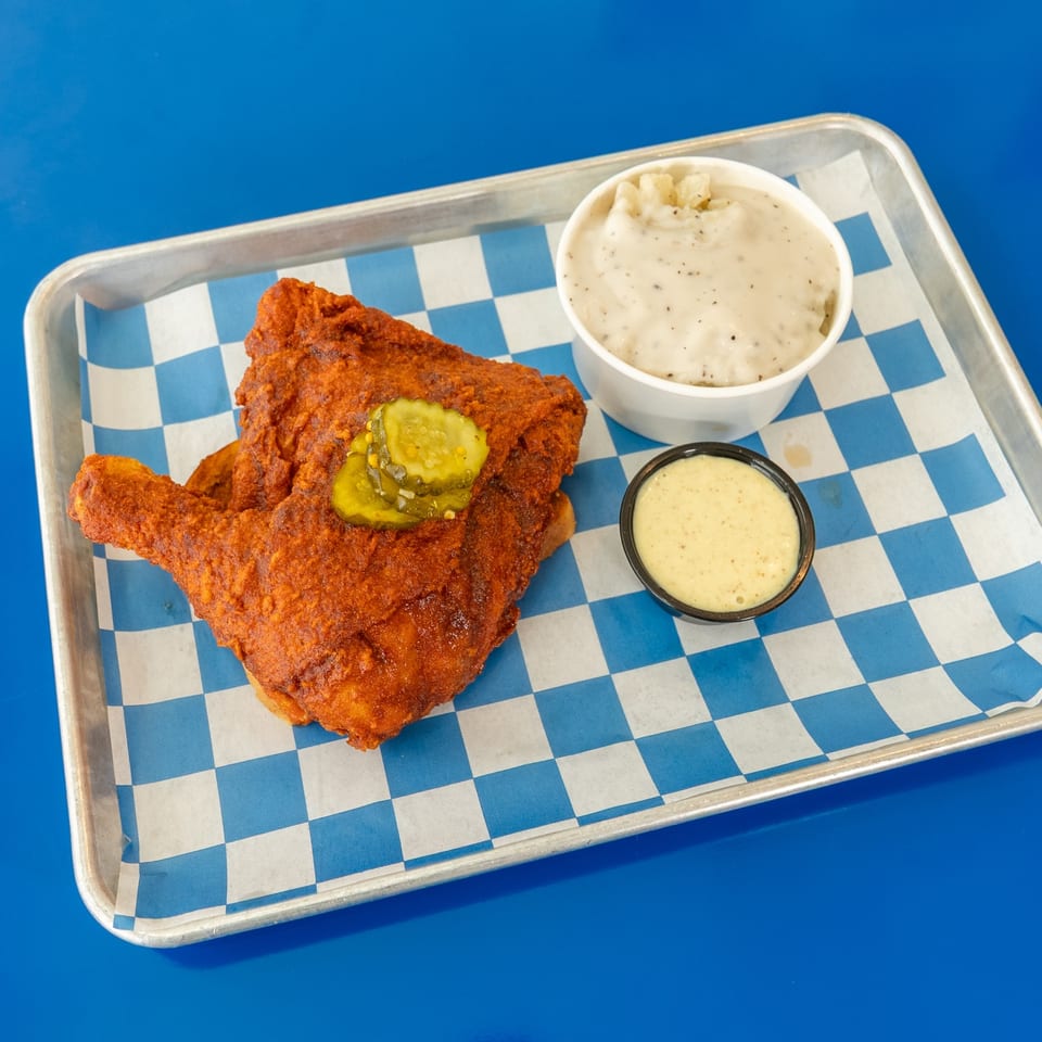 Crispy fried chicken tenders topped with pickles, served with mashed potatoes and dipping sauce on a checkered tray.