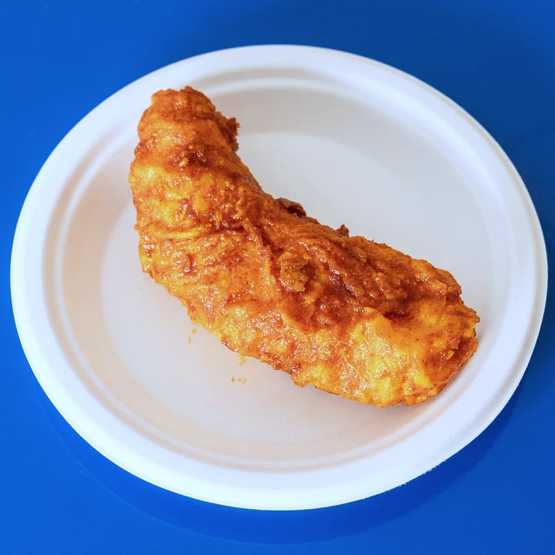 A golden-brown fried fish fillet rests on a white paper plate against a blue background.