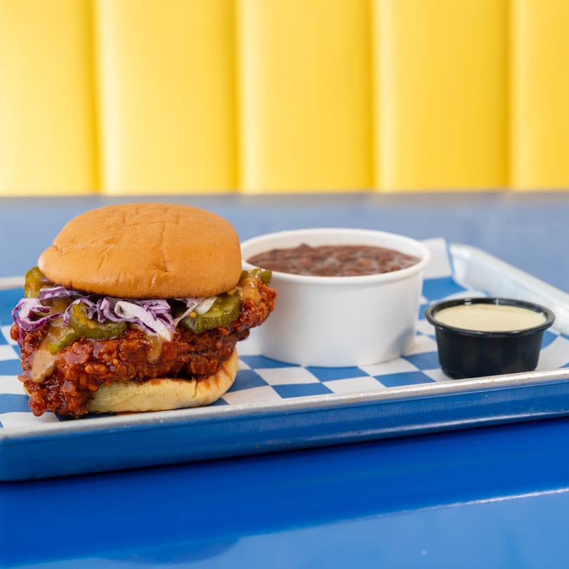 Spicy chicken sandwich with coleslaw and pickles, side of beans and sauce on a blue tray.