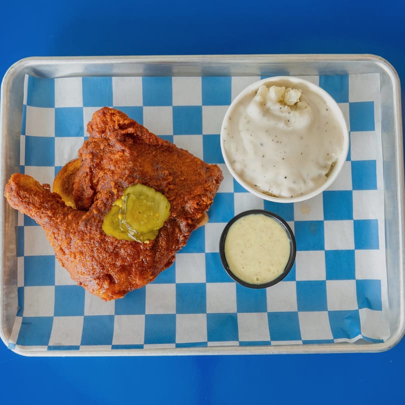 Crispy fried chicken with pickles, mashed potatoes, and dipping sauce on a checkered tray.