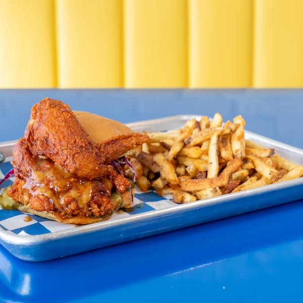 Crispy fried chicken sandwich with sauce and pickles, served with seasoned fries on a metal tray.