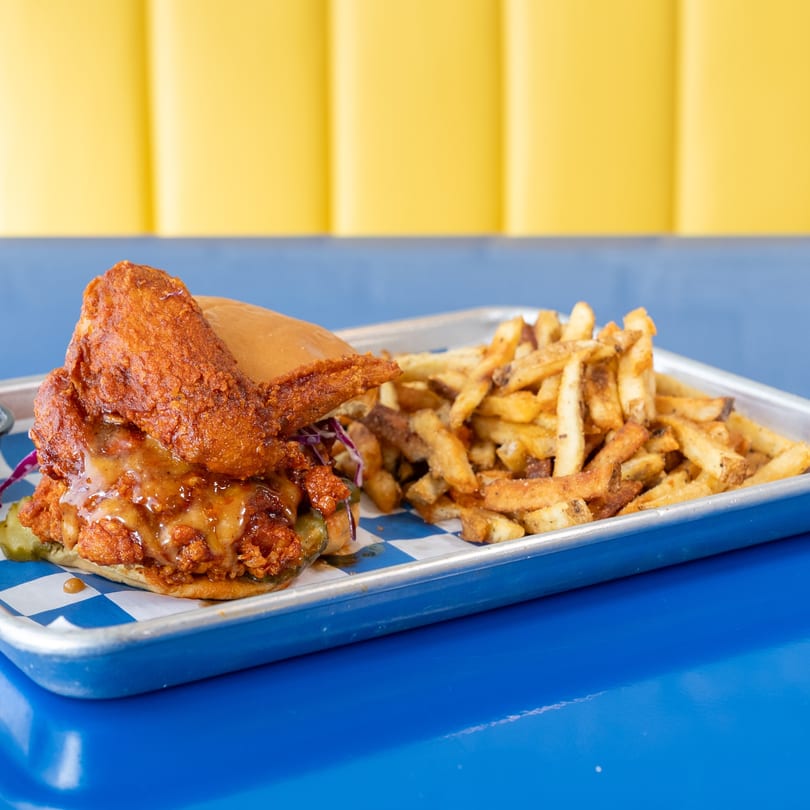 Crispy fried chicken sandwich with sauce and pickles, served with seasoned fries on a metal tray.