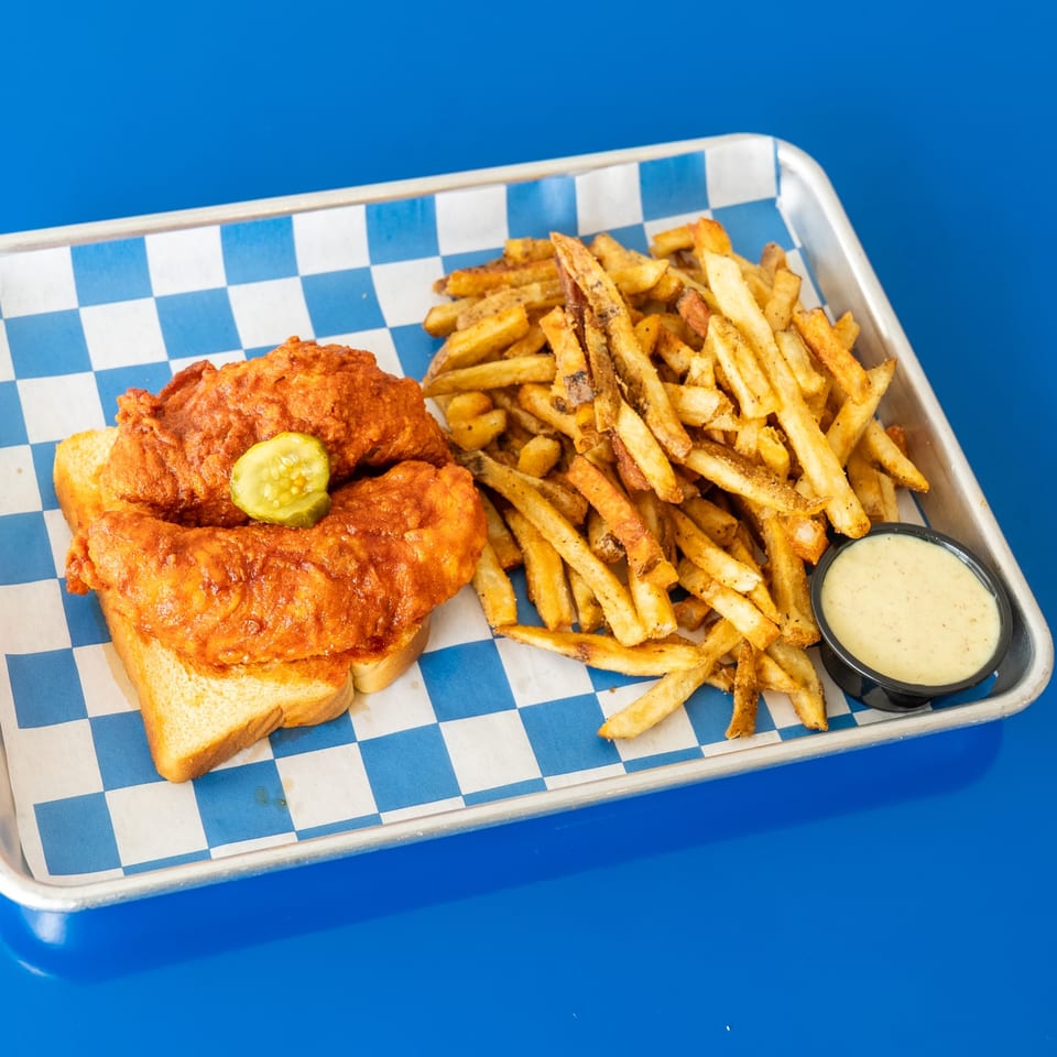 Crispy fried chicken sandwich with a pickle slice, seasoned fries, and dipping sauce on a blue-and-white checkered tray.
