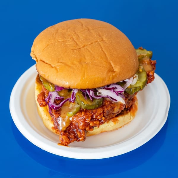 Loaded burger with a golden bun, saucy fried filling, pickles, purple cabbage, onions, and cheese on a white plate.