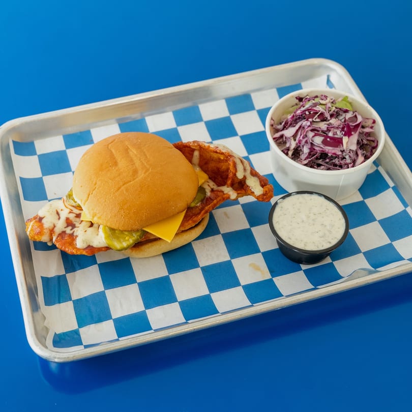 Spicy chicken sandwich with cheese and pickles, coleslaw, and ranch dressing on a blue checkered tray.