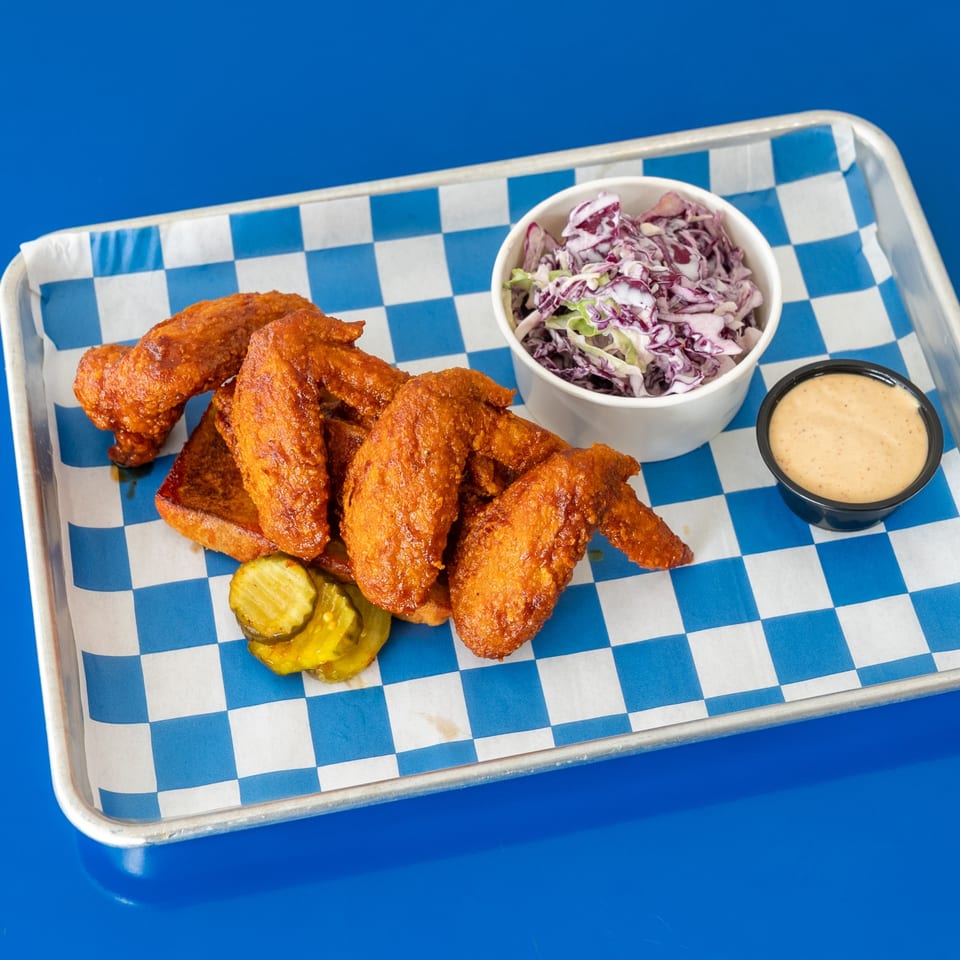 Crispy fried chicken wings with toast, pickles, purple coleslaw, and dipping sauce on a blue-and-white checkered tray.